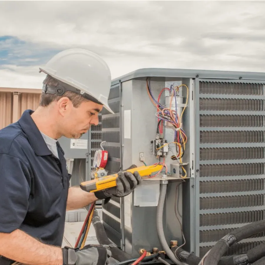 HVAC technician in a hard hat testing an outdoor air conditioner with a multimeter